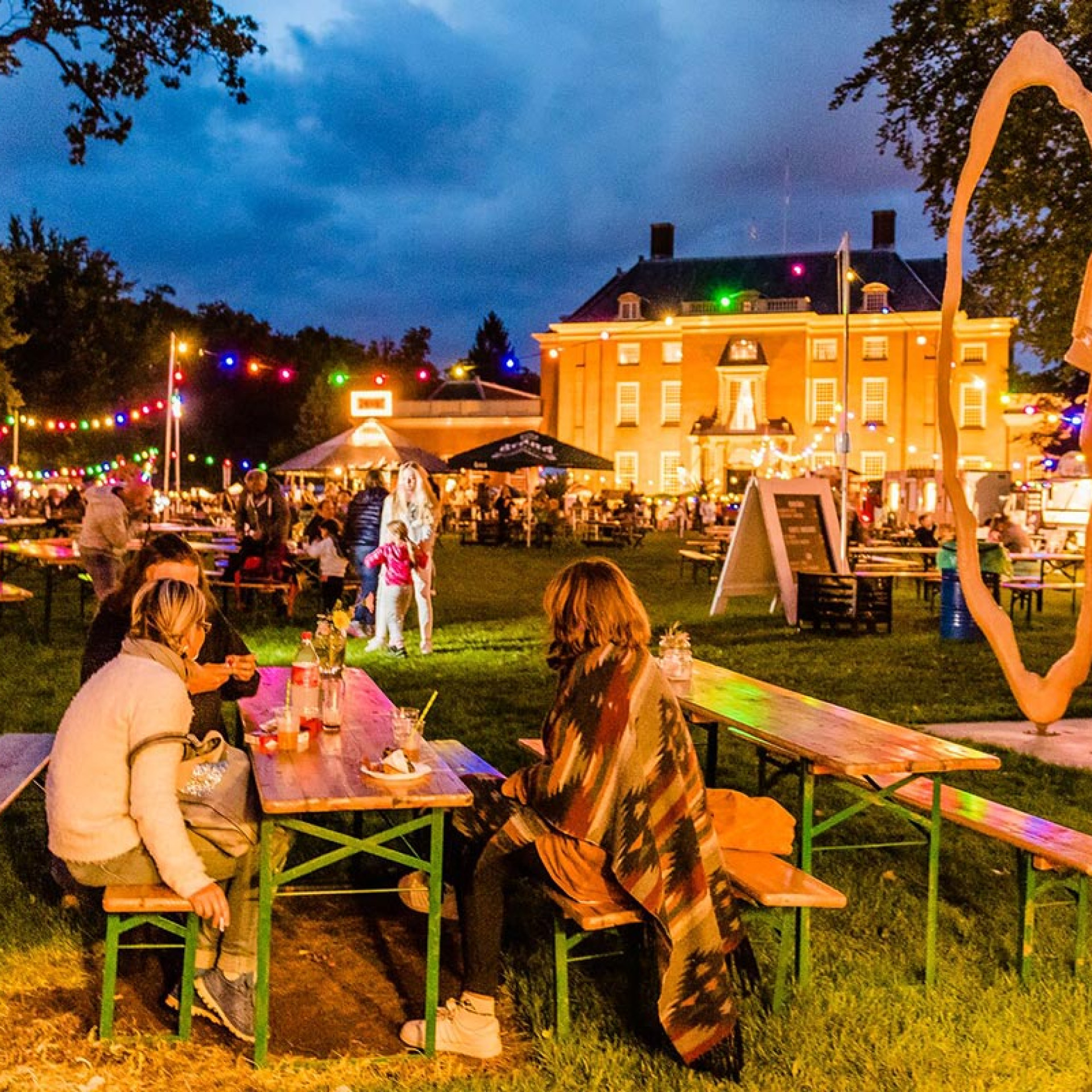 Food Festival in de tuin van Slot Zeist. Mensen genieten van hapjes en drankjes aan tafels, omgeven door de prachtige tuinen van het kasteel.